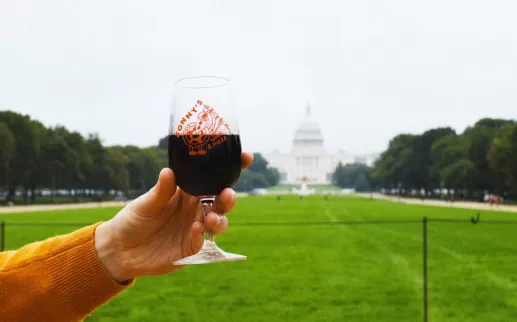 A hand raises a glass of red wine on the National Mall with the U.S. Capitol in the background.
