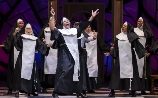 A nun holding a conductor ’ s wand dances animatedly with arms stretched up in the air as a large group of nuns dance and sing behind her.
