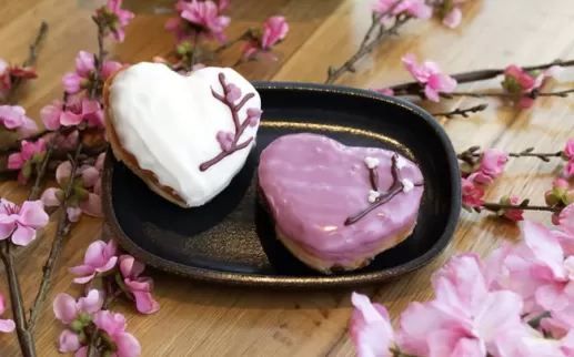 Two heart-shaped donuts decorated with cherry blossom branches sit on a table staged with cherry blossoms. 