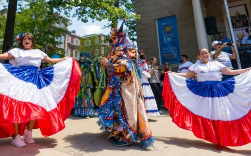 Two women in colorful skirts dance alongside a costumed figure, celebrating embassies in Washington, DC. 