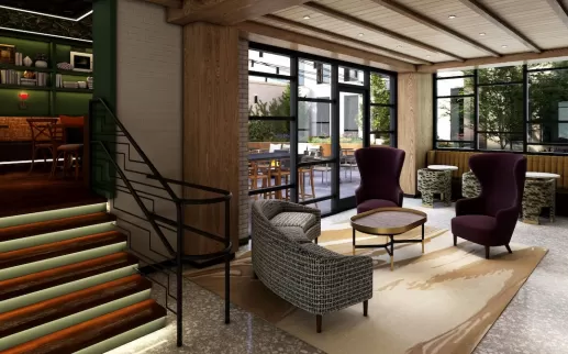 A stylish hotel lobby featuring a mix of modern and classic furnishings, with cozy lounge chairs, a carpeted seating area, a staircase leading to a book-lined nook and large windows letting in natural light.