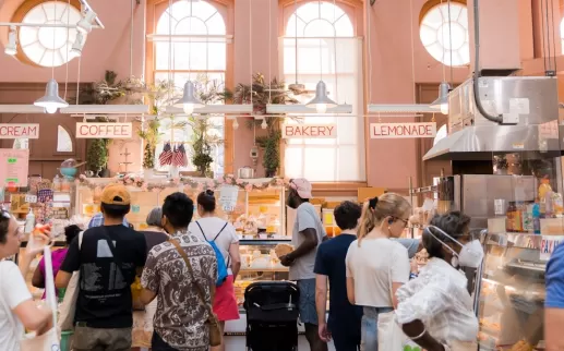 Shoppers queue at a brightly lit indoor bakery and deli inside Eastern Market, with signs for sandwiches, coffee and lemonade.