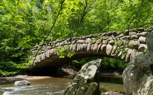 A picturesque stone bridge arches over a gently flowing creek surrounded by lush green forest in spring.