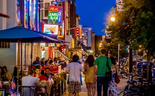 People walk down a street lit up with neon signs and busy restaurants and bars. 
