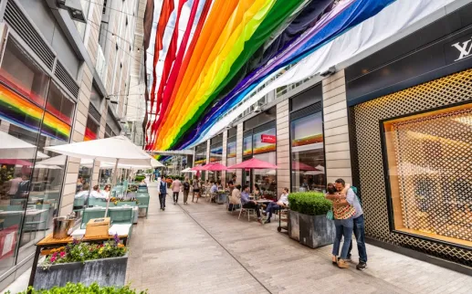 Colorful rainbow fabric banners hang above a lively pedestrian alley at CityCenterDC, where people dine outdoors and stroll between modern shops and restaurants.