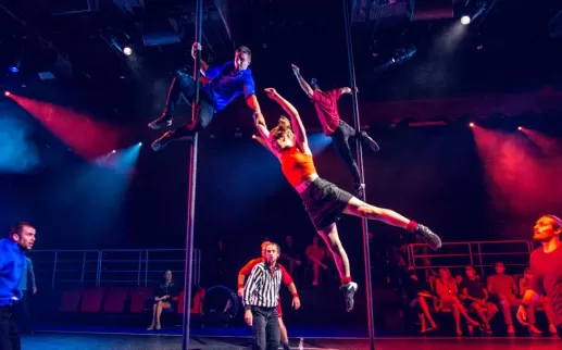 Acrobats perform daring moves on poles while a referee and audience watch during a high-energy scene from Duel Reality.
