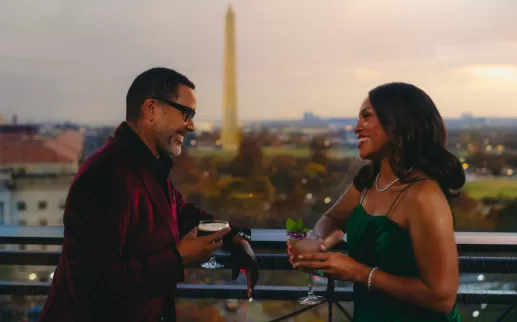 A well-dressed couple enjoys cocktails at The Vue rooftop restaurant with the Washington Monument in the background at sunset in Washington, DC.