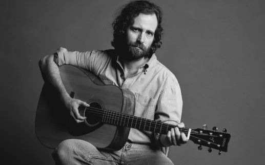 Comedian and musician Kyle Mooney poses with an acoustic guitar in a black and white promotional portrait.
