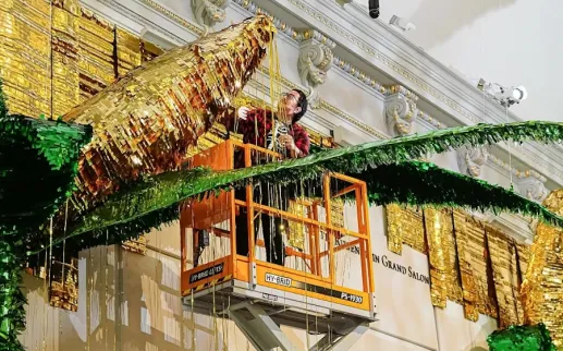Artist installs a large gold and green piñata-style artwork from a lift inside a grand gallery.
