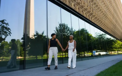 A couple holds hands while walking outside the Smithsonian National Museum of African American History and Culture, with the Washington Monument reflected in the glass.