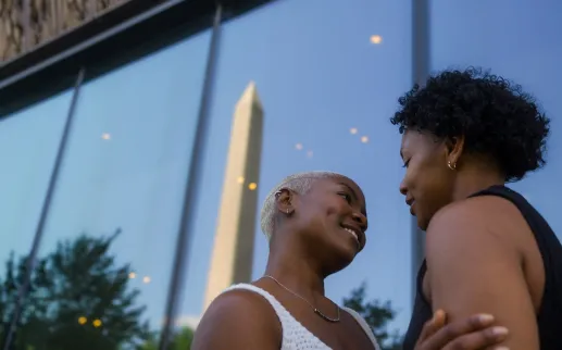 A couple shares an intimate moment in front of the Smithsonian National Museum of African American History and Culture, with the Washington Monument reflected in the glass.
