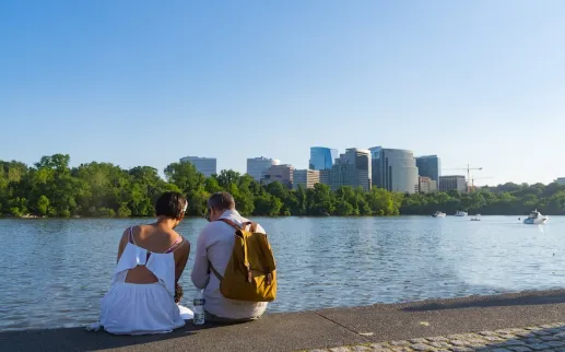 A couple sits by the Georgetown waterfront, looking out across the Potomac River toward the skyline of Rosslyn, Virginia.