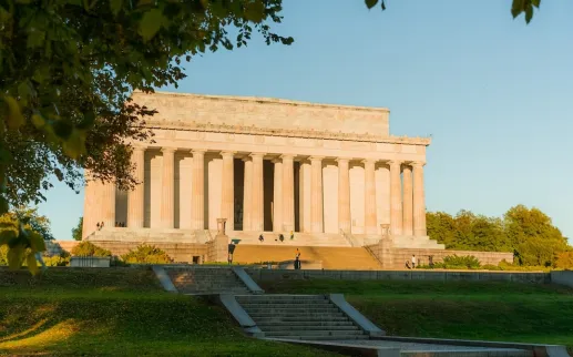 The Lincoln Memorial at sunset framed by trees with golden light on the monument’s columns.
