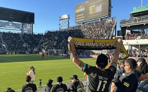 A Washington Spirit fan holds up a yellow scarf reading “Washington, DC” in a packed Audi Field stadium during a soccer match.
