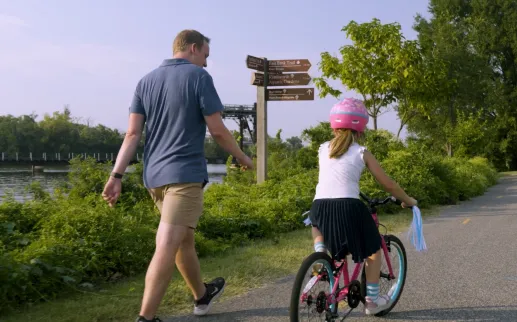 A man walks beside a young girl riding a bike along a riverside trail surrounded by greenery in Washington, DC.