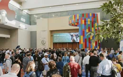 A large crowd of conference attendees gathers in a bright, spacious convention center lobby with colorful signage and modern architectural elements.
