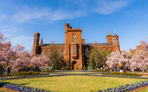 Visitors walk across the National Mall toward the red sandstone Smithsonian Castle on a sunny day.