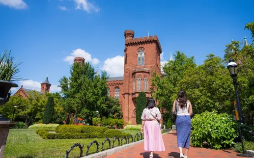 Two women walk along a garden path toward the Smithsonian Castle surrounded by lush greenery.