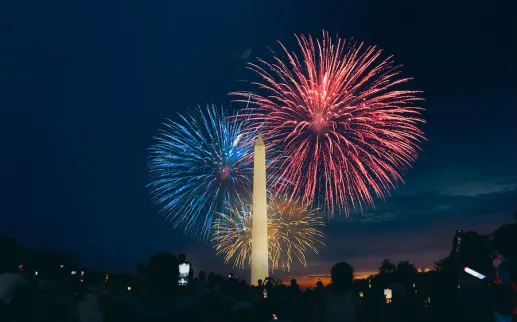 Fireworks burst in red and blue above the Washington Monument as crowds watch on the National Mall at night.
