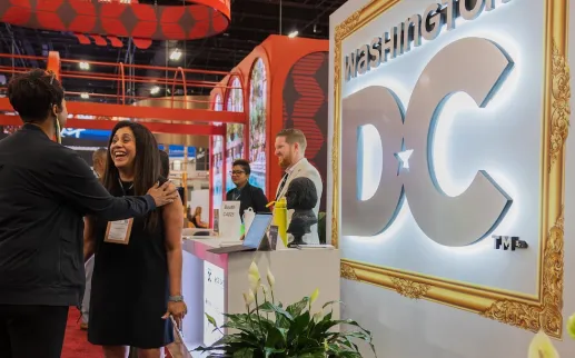 Attendees gather around a Washington, DC destination booth featuring illuminated signage and large digital displays at a travel trade show.
