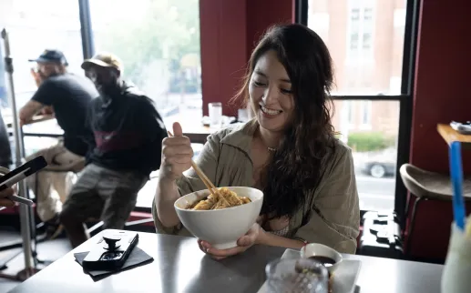 A guest smiles while enjoying a bowl of noodles at the bar inside Chaplin’s Restaurant in Washington, DC, with other diners and daylight visible through the windows.