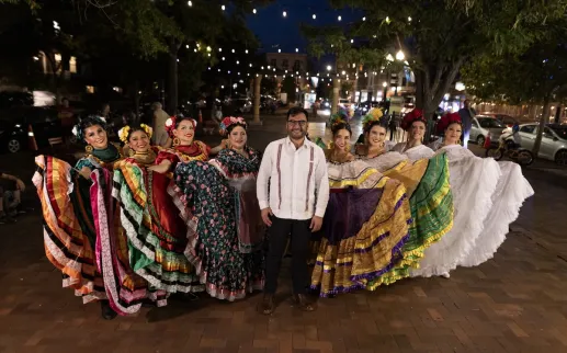 A group of folklórico dancers in colorful traditional dresses pose together outdoors at night during a cultural performance in Washington, DC.
