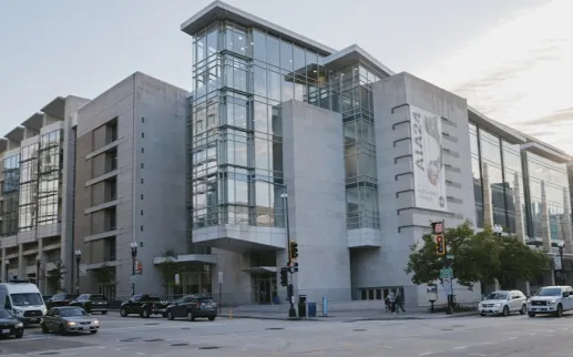 A modern glass-and-concrete museum building on a city corner with cars and pedestrians under a partly cloudy sky.