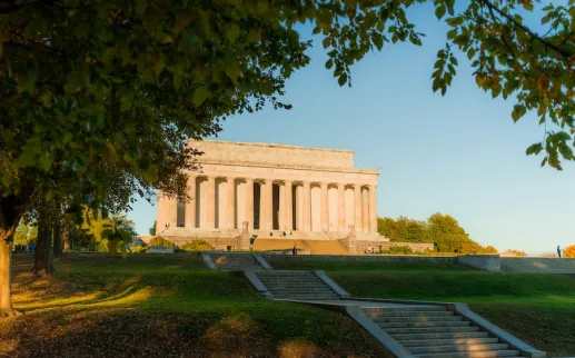 The Lincoln Memorial is seen from a distance, framed by leafy trees and lit by warm sunlight.