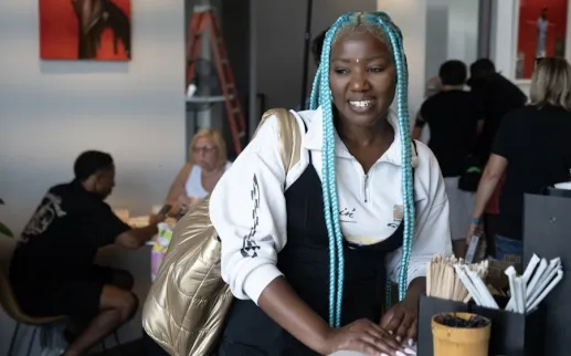 A person with long blue braids smiles while leaning on a café counter with people seated in the background.
