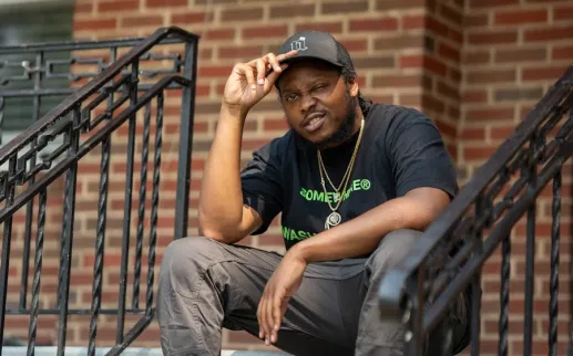 A man in a black T-shirt and cap sits on the steps of a brick building, looking toward the camera.