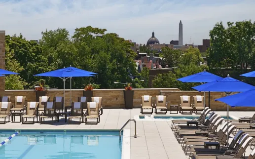 Rooftop pool deck with lounge chairs and blue umbrellas overlooking a city skyline