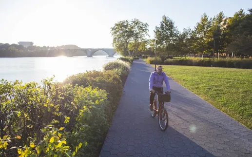 Cyclist on a riverside path at sunset, with trees and green grass nearby