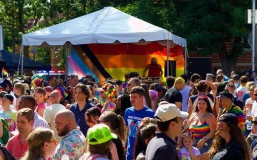 Crowds celebrating at the 17th Street Pride Block Party, gathered around a DJ booth beneath a rainbow Pride flag backdrop, with attendees wearing colorful outfits and accessories.