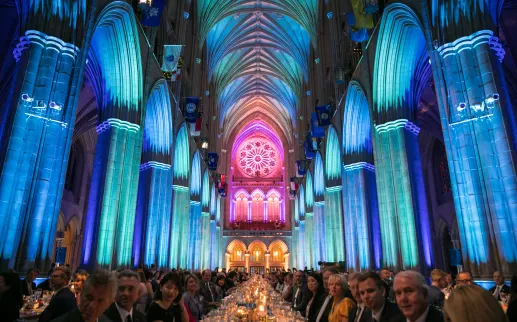 The soaring Gothic arches and vaulted ceiling of Washington National Cathedral are illuminated in vibrant blue, green and pink lighting, highlighting the ornate rose window at the far end of the nave
