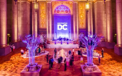**Alt text:** Guests gather beneath dramatic purple lighting in the ornate Andrew W. Mellon Auditorium for a reception featuring illuminated cocktail tables and a view toward the stage.