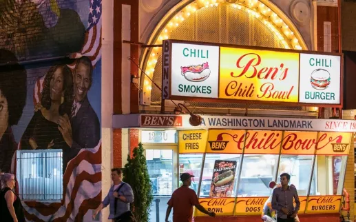 Nighttime street scene with bright restaurant signs and people inside behind glass windows
