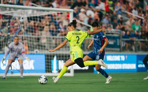Photo of soccer player in yellow kicking toward goal, defended by blue-uniform player, packed crowd
