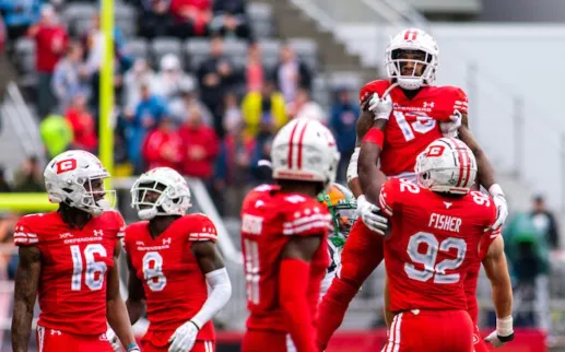 Photo, football players in red uniforms celebrate on the field as crowd watches.