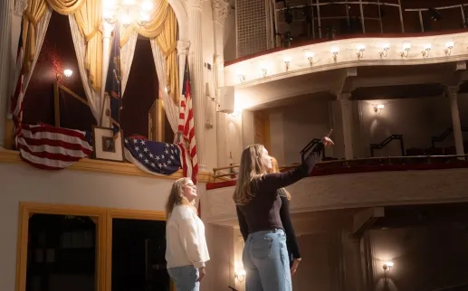 Two performers on stage beneath ornate theater balconies and chandelier.