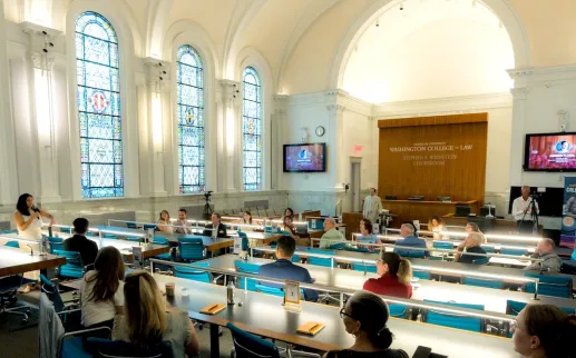 Photo of people at long tables in a bright hall with arched windows and a projector screen
