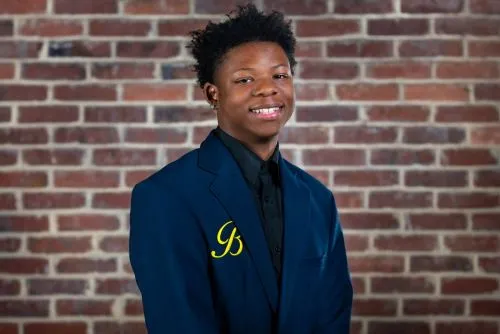 John "JT" Thompson, un joven negro, se encuentra frente a un fondo de ladrillo. Lleva una chaqueta azul marino con el escudo de la Academia de Hotelería y Turismo de Ballou.