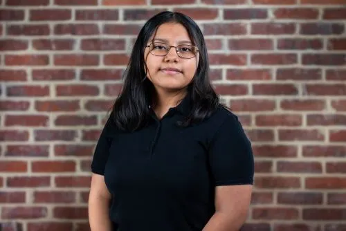 Melany Méndez Casas, una joven latina, parada frente a una pared de ladrillos. Lleva gafas y un polo negro.