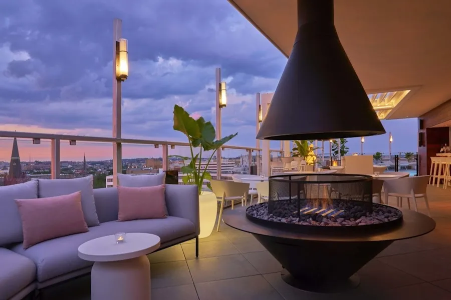 Rooftop lounge at sunset with modern furniture, including a sectional sofa with pink pillows, round tables, and a central fire pit. The area is illuminated with contemporary lighting and offers panoramic city views.