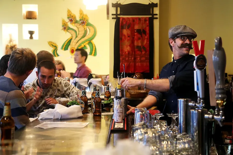 Bartender with a mustache serves drinks to patrons at a busy bar, decorated with vibrant artwork in the background.
