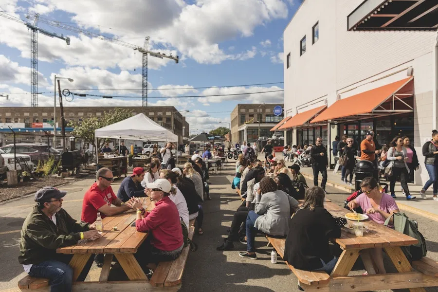 Groups of people enjoying food and drinks at outdoor picnic tables on a sunny day.