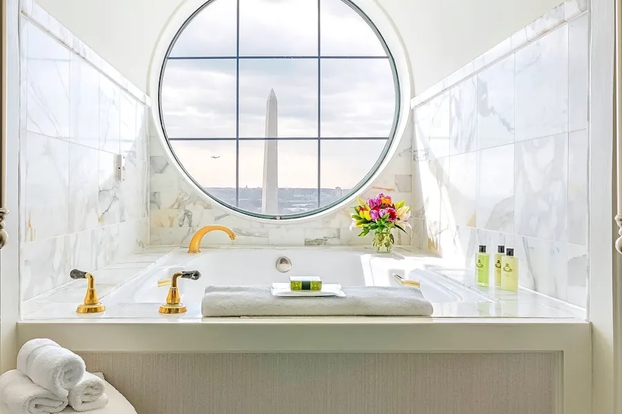 A luxurious bathroom with a marble bathtub and golden fixtures, featuring a round window with a view of the Washington Monument. The bathtub has towels, flowers, and toiletries arranged neatly.