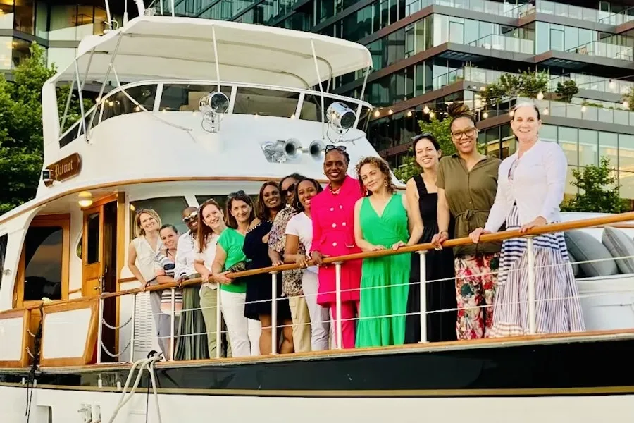 Um grupo de pessoas em pé ao longo da grade de um iate, sorrindo para uma foto de grupo. O iate está atracado, com um moderno edifício de vidro ao fundo, refletindo a luz do entardecer. O grupo está vestido com uma mistura de trajes casuais e semiformais, criando um clima de celebração.