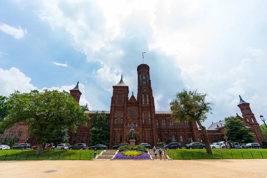 Vista externa do Castelo Smithsonian no National Mall, com uma estátua de Joseph Henry na frente e pessoas caminhando nas proximidades sob um céu parcialmente nublado.