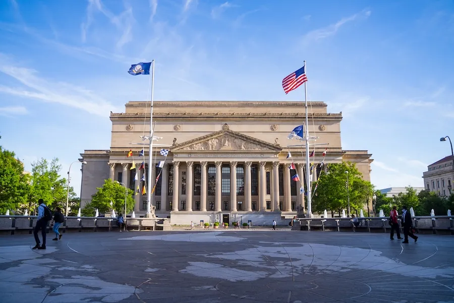  The National Archives building stands under a clear blue sky, framed by two flagpoles with American and military flags at the entrance, and a few people walking in the foreground.