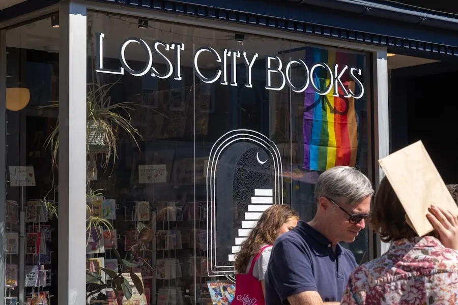 An exterior shot of Lost City Books in the daytime, with books and a rainbow flag visible inside. 